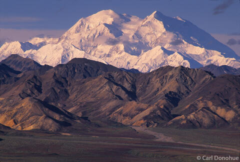 The snow-covered summit of Denali, North America's highest mountain, dominates the horizon above a series of rust-colored autumn foothills and a braided river.