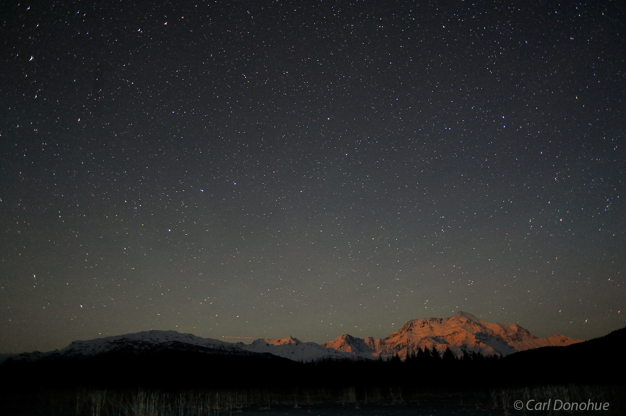 A starry night sky falls above Mt. Saint Elias, still glowing in the late evening sun. Stars at night over Mt. St. Elias, Icy...
