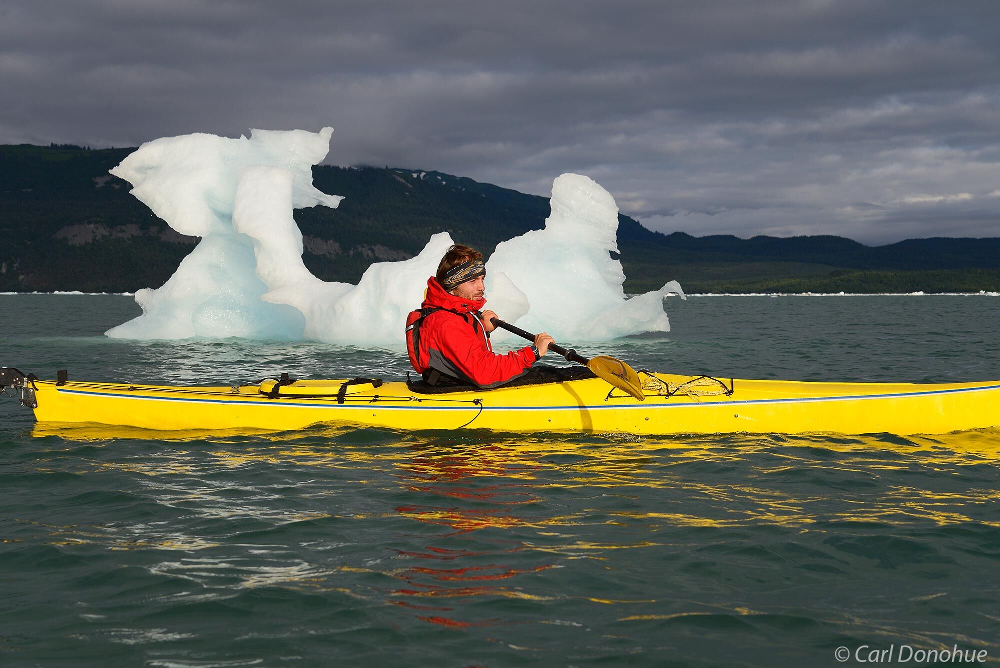 A stock photo of a young man sea kayaking past a large iceberg in Icy Bay, with icebergs floating in the background as he paddles...
