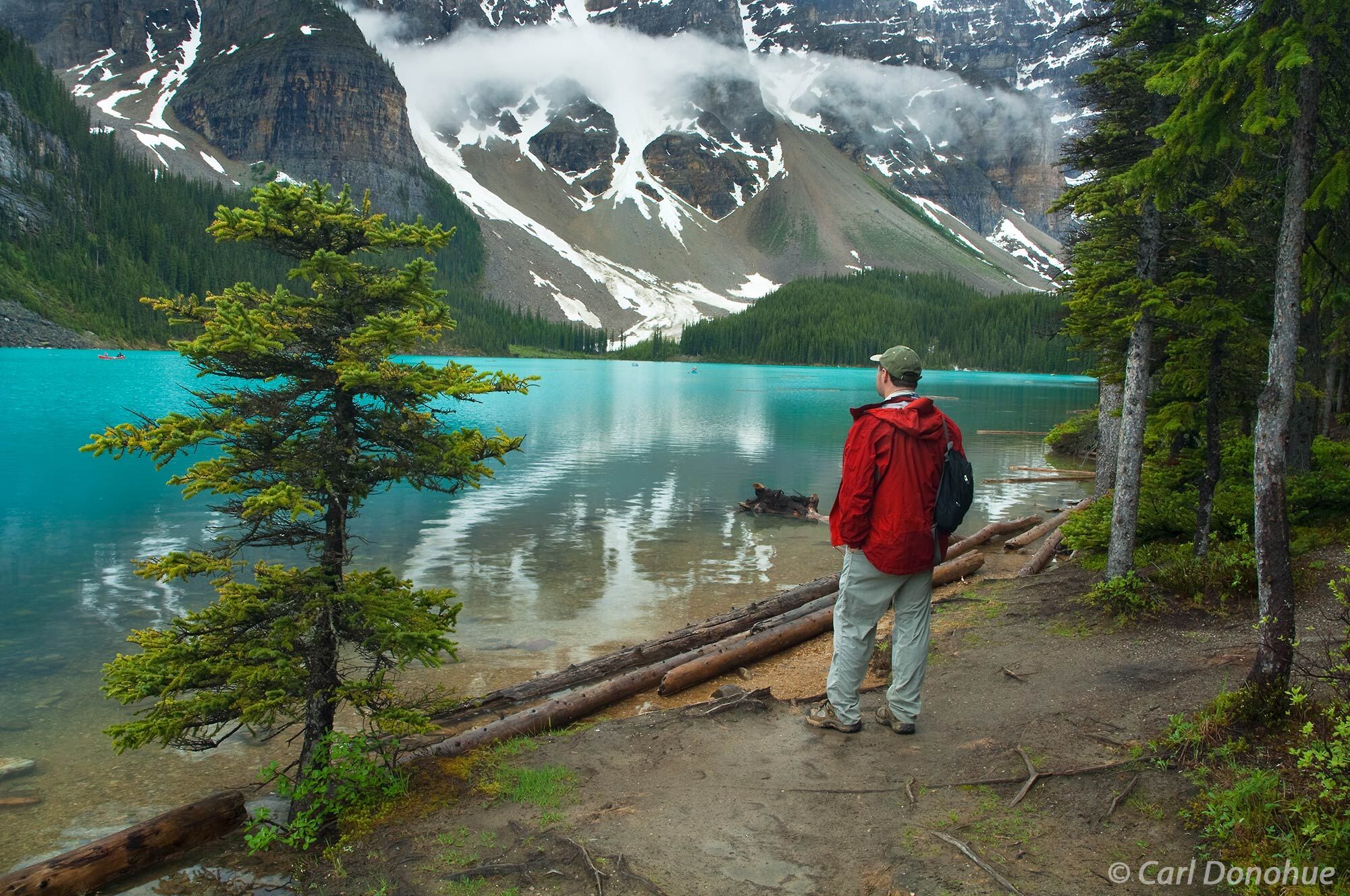 A tourist hiker stands beside the shores of Moraine Lake and watches people canoeing on the lake The grand scenery of Moraine...